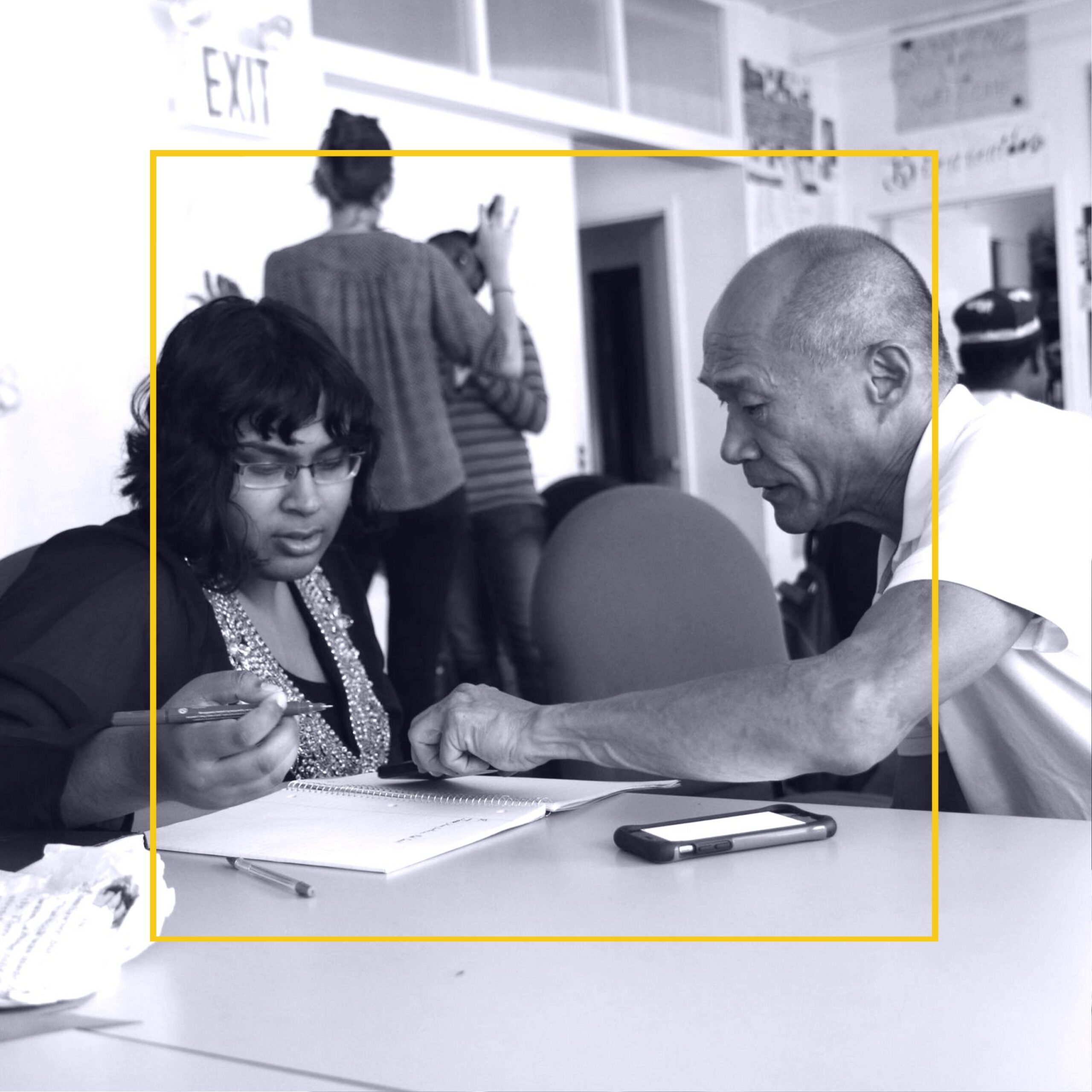 Black and white photo of older balding Asian man mentoring young short, dark haired southeast Asian woman holding a pen. They both are looking at a notebook on a table.