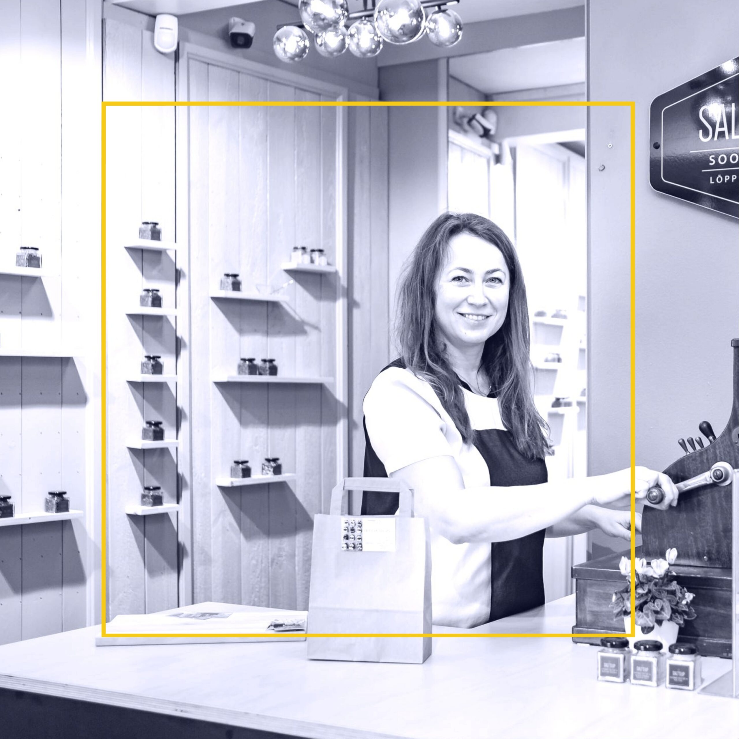 Black and white photo of white woman smiling and standing in front of old time cash register and counter with bag on the counter. Items line the shelves on the back walls of the shop.