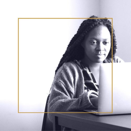 Black and white photo of Black woman with long braided hair sitting at a laptop with a slight smile on her face looking pleased.